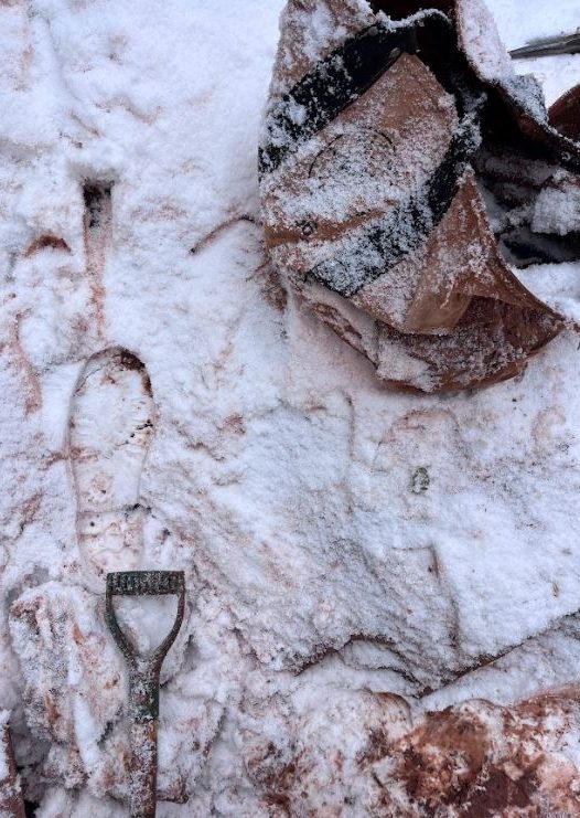Snowy ground, rock bag lightly dusted in snow in upper right corner, shovel handle in lower left corner lightly dusted in snow