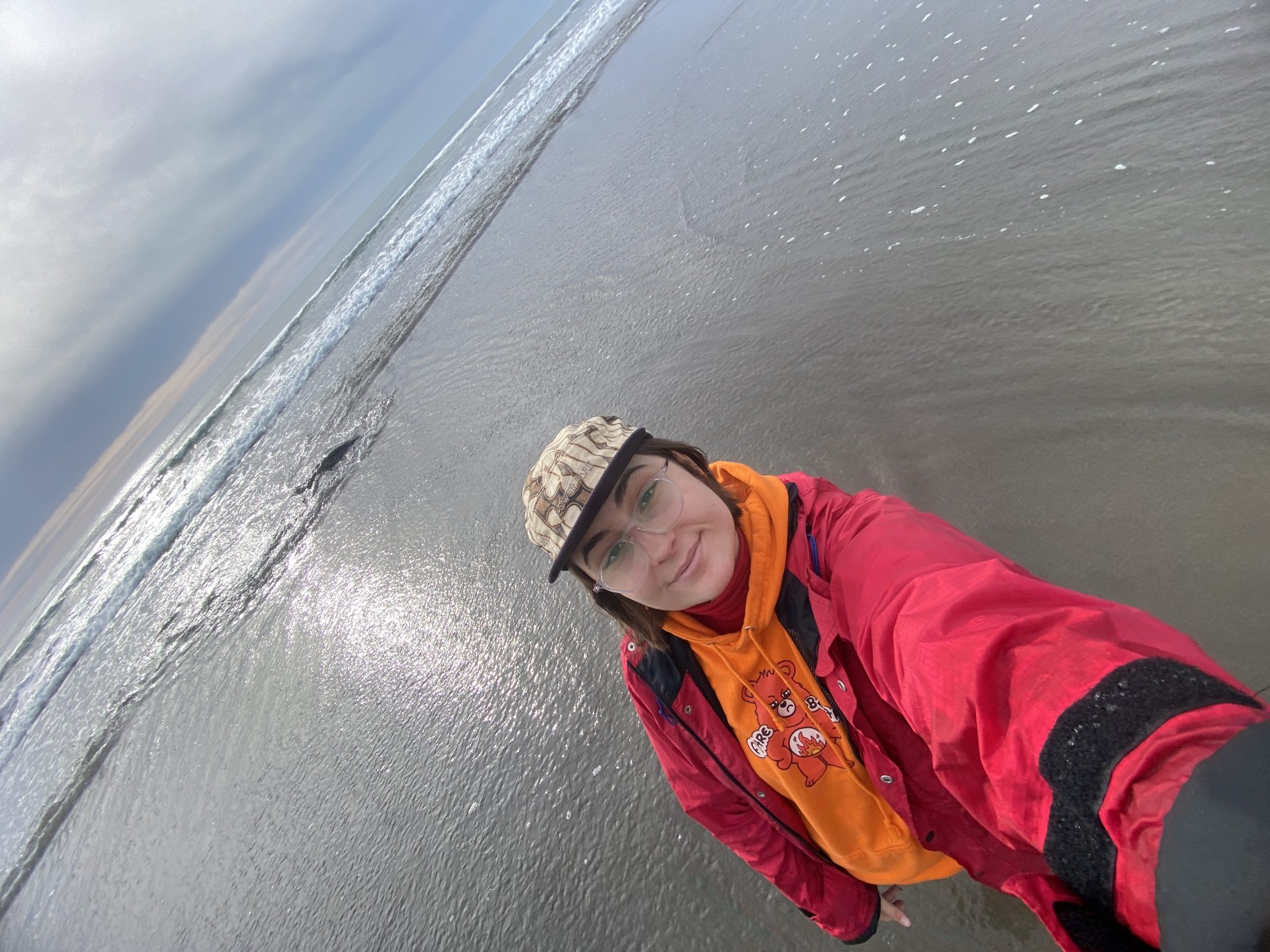 A 0.5 selfie of Mo, a white non-binary smiling individual with short brown hair. They wear clear glasses, a brown and tan cap, a neon orange sweatshirt, and a red windbreaker. They stand on the beach at the edge of the water. Small waves are in the background.