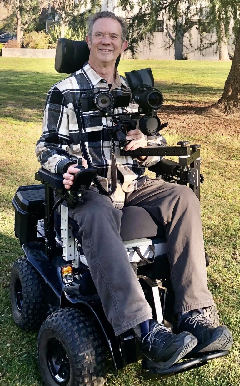 An older man with short grey hair, a white and black plaid shirt and brown pants smiles at the camera. He is a power wheelchair user and has adaptable binoculars and a camera attached to the armrest of his chair. His wheelchair features off-terrain wheels. He is on grass and a tree is behind him.