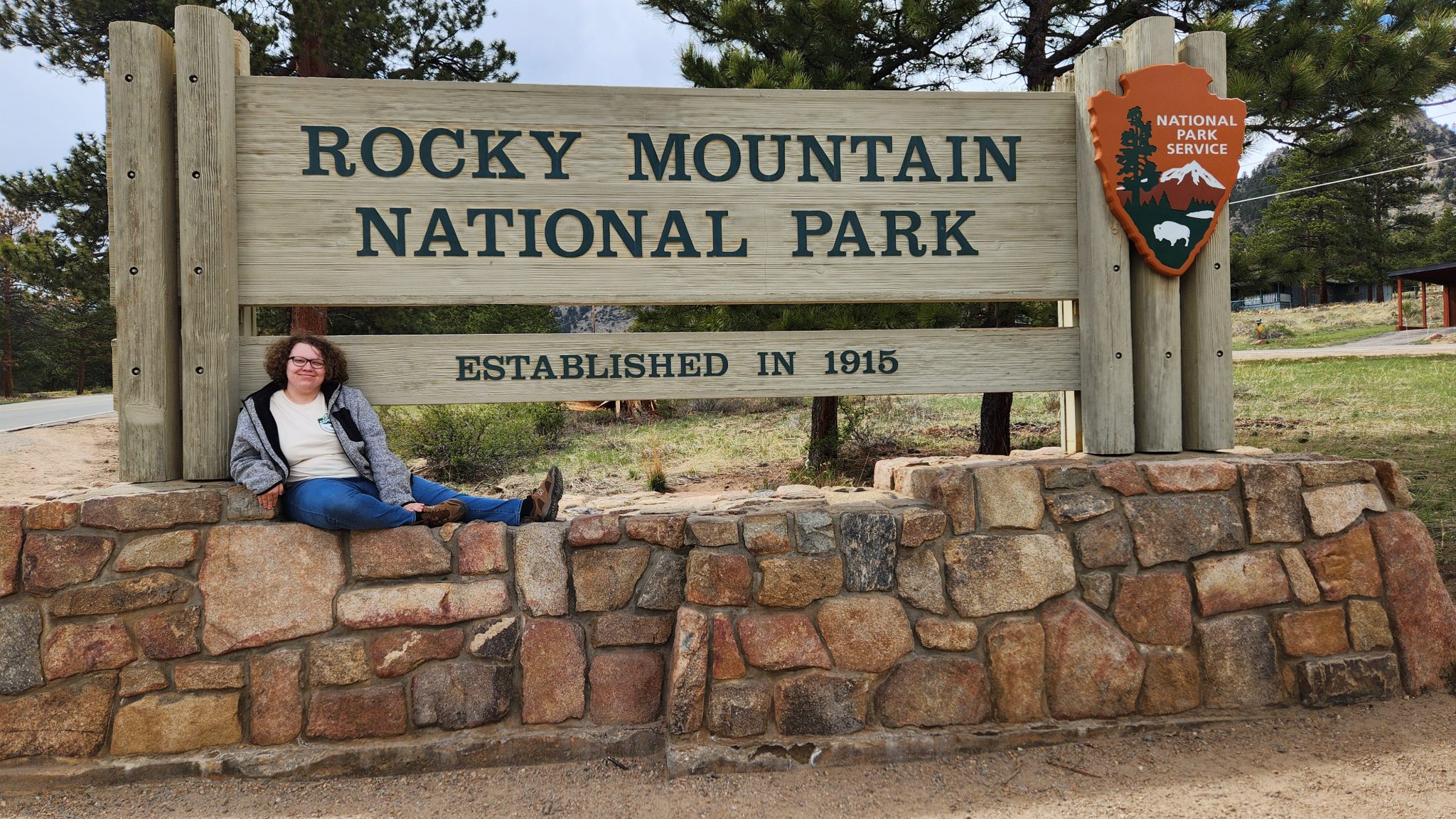 A women in her twenties sitting to the left of the Rocky Mountain National Park sign.