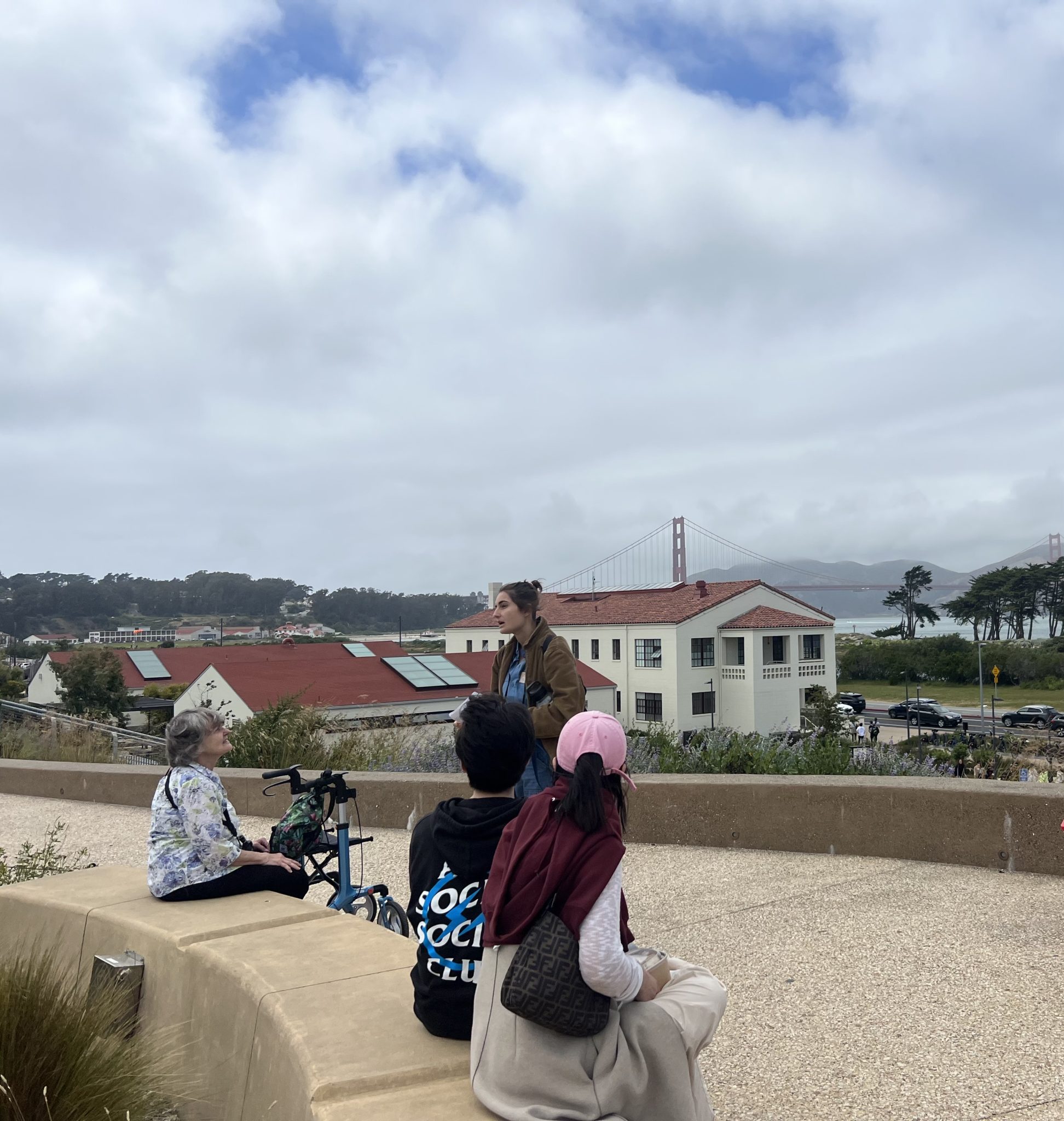 Three people sit on concrete seating looking at someone who is speaking; one person uses a walking aid. In the foreground is a wide trail the group is on. In the background are white and red buildings, grass, trees and the Golden Gate Bridge.