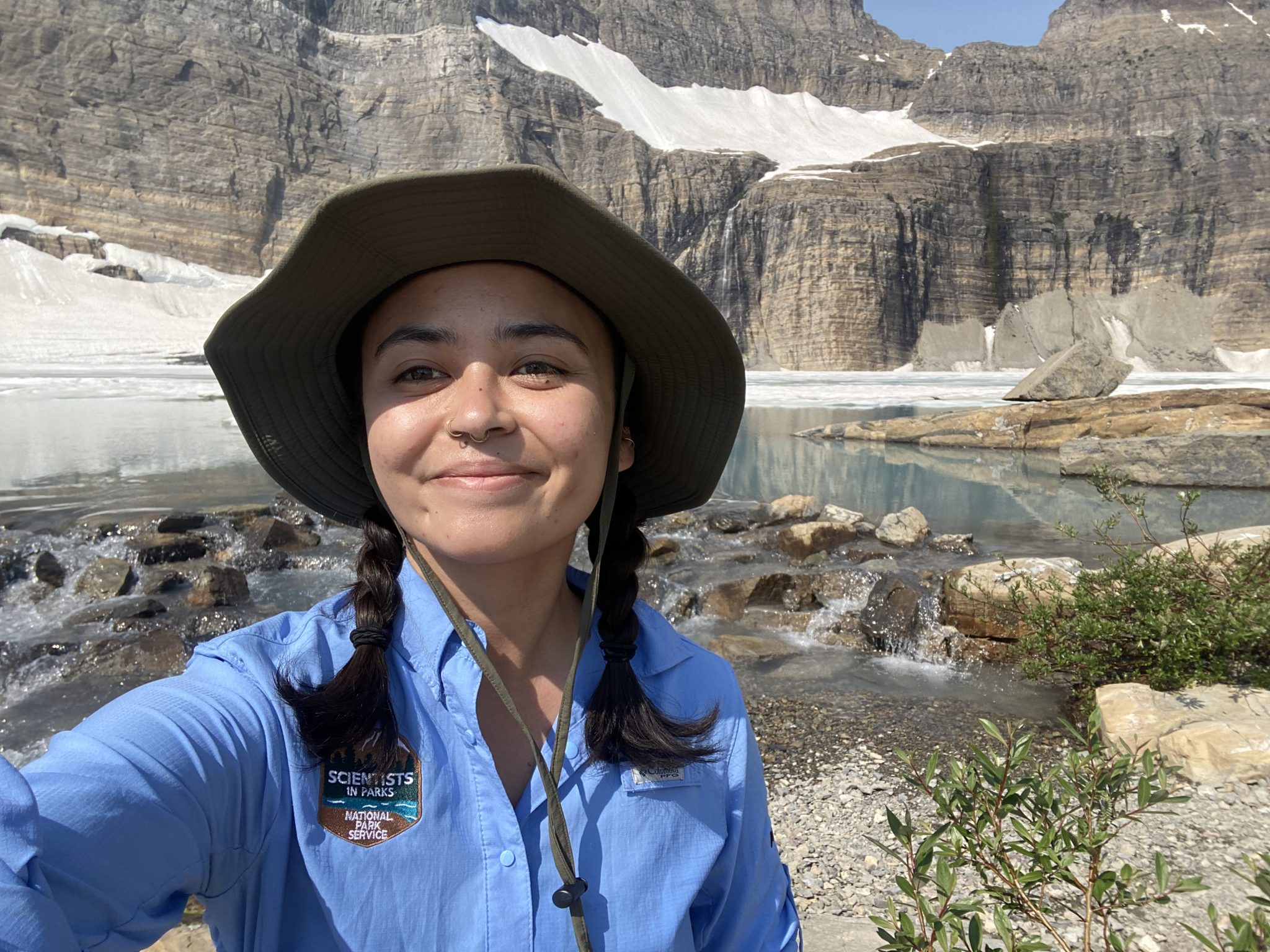 Sophia Britto, a Mosaics in Science intern, stands in front of Grinnell Glacier, wearing a blue MPS shirt and hat.
