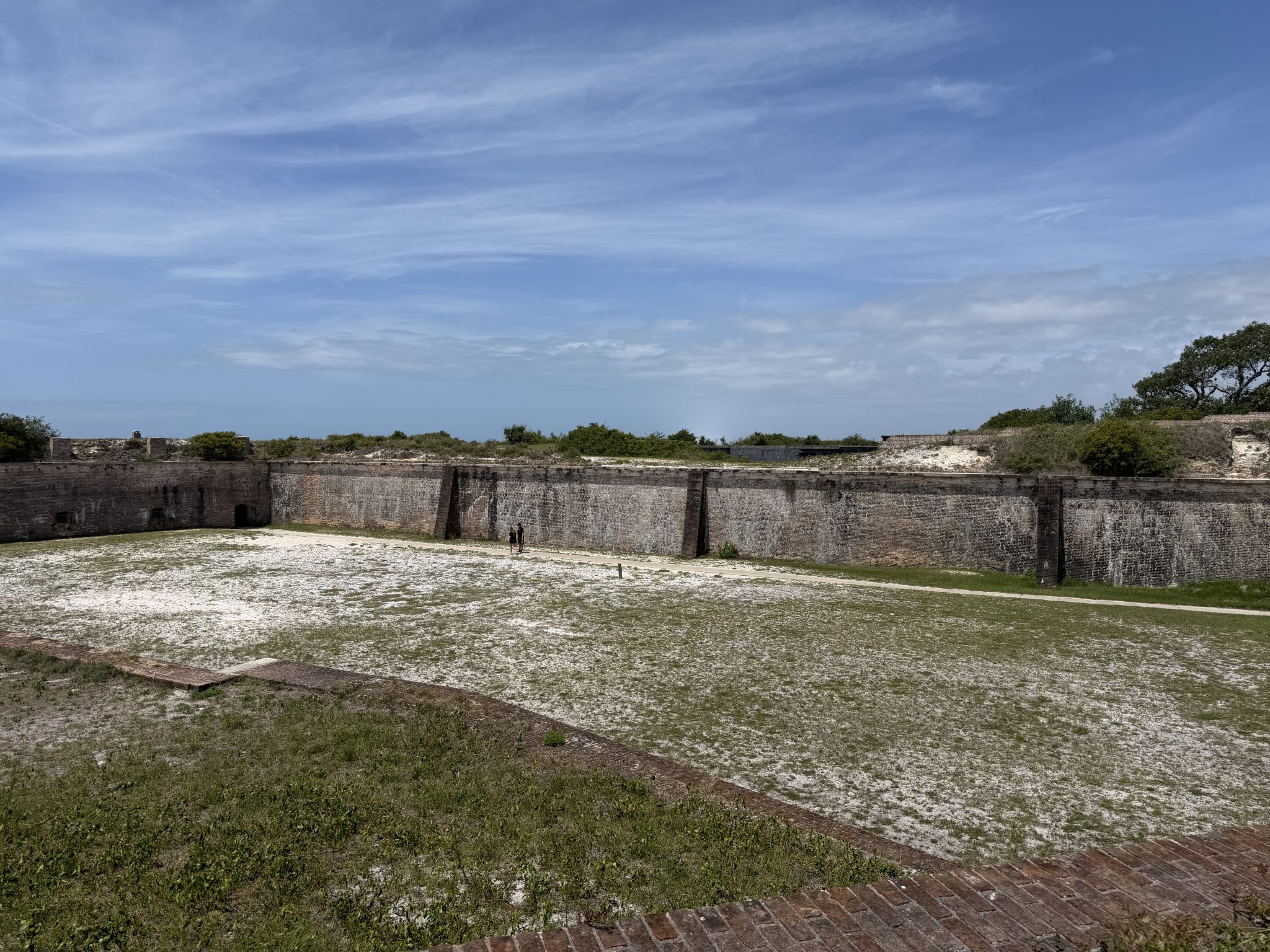 Wide view inside Fort Pickens showing a large open sandy courtyard surrounded by tall historic brick fort walls under a bright blue sky with wispy clouds.