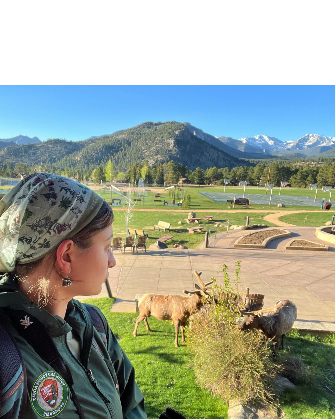 A young person wearing a floral headscarf and a green jacket with a National Park Service Youth and Young Adult Programs patch looks out at two elk grazing on a grassy lawn. Behind them is a landscaped courtyard with benches, forested foothills, and snow-capped Rocky Mountain peaks under a clear blue sky.