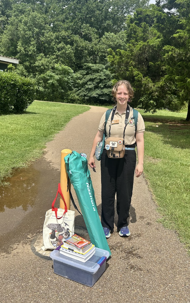 AG with binoculars and a backpack on, standing in front of a pole marking the entrance to a trail. By the pole, there is a bin of binoculars, a World Migratory Bird Day tote bag, two folded trail chairs in their bags, and a stack of field guide books.