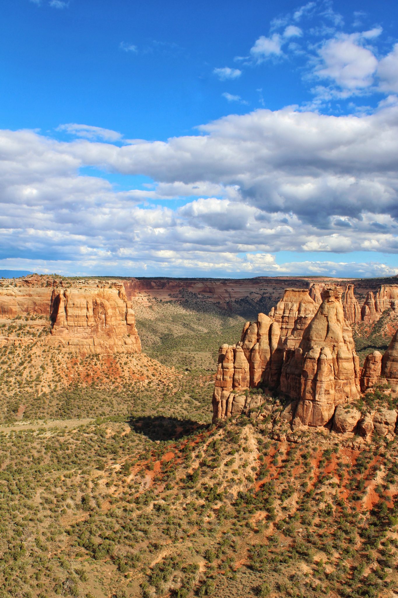 Towering orange sandstone monoliths jut out from canyon walls. The monolith on the left has numerous smaller spirals, The canyons stretch to the horizon, and the deep canyon floors are covered in green shrubbery. The sky is a bright blue with partial cloud cover, and the clouds look like they are on the verge of becoming rain clouds.