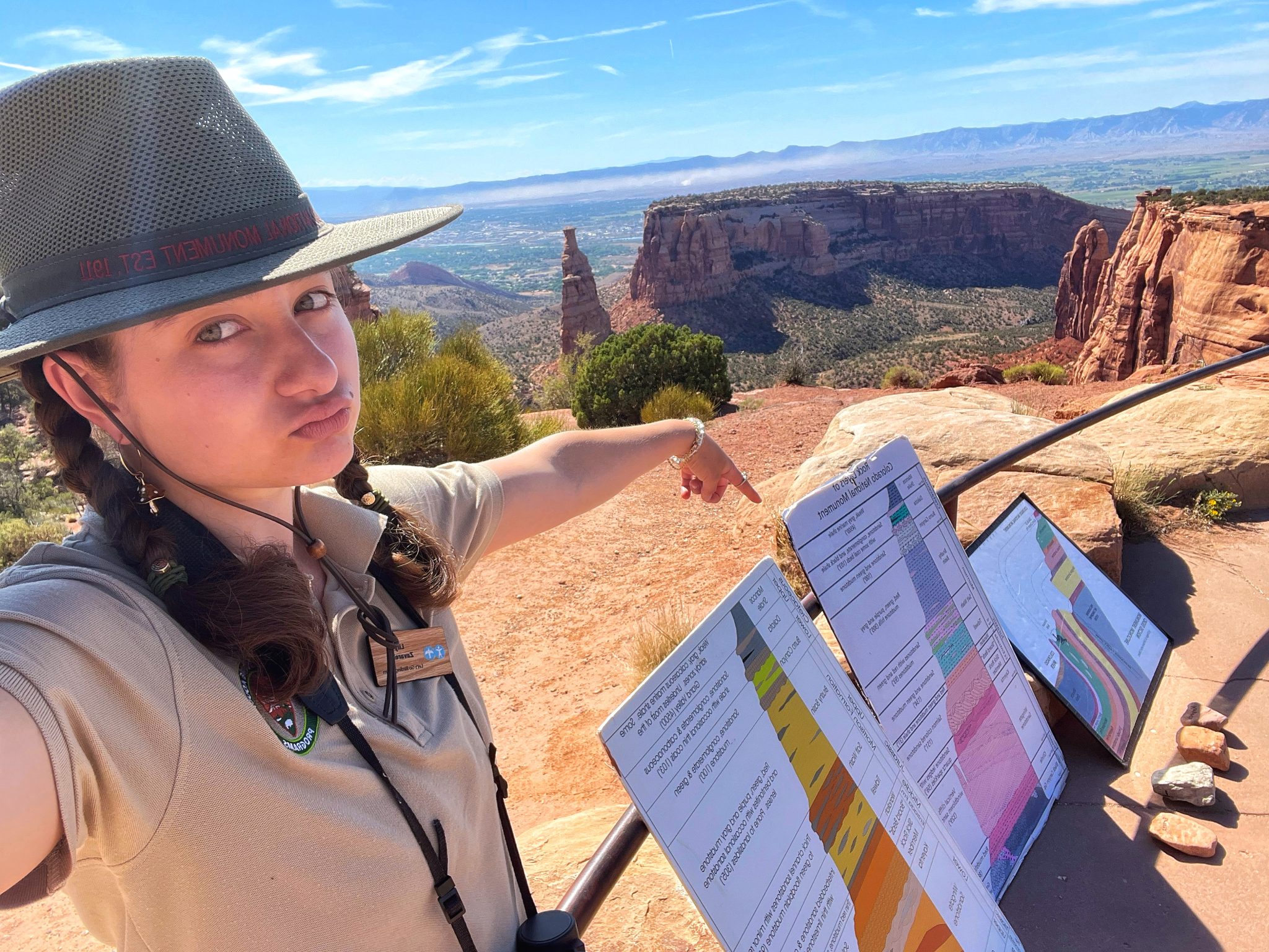 Lily takes a selfie with her educational geology material. Lily is on the left side of the picture, wearing a beige collared t-shirt with the NPS arrowhead logo and her wooden nametag across her chest. Her brown hair is in two pigtail braids, and atop her head is a green flat-top ranger hat. Her left arm is extended to point at three posters displaying the colorful rock layers of the Monument. The posters are propped up against a railing, and behind is a beautiful red rock canyon view covered by a scrubby pinyon-juniper woodland under an almost clear blue sky.