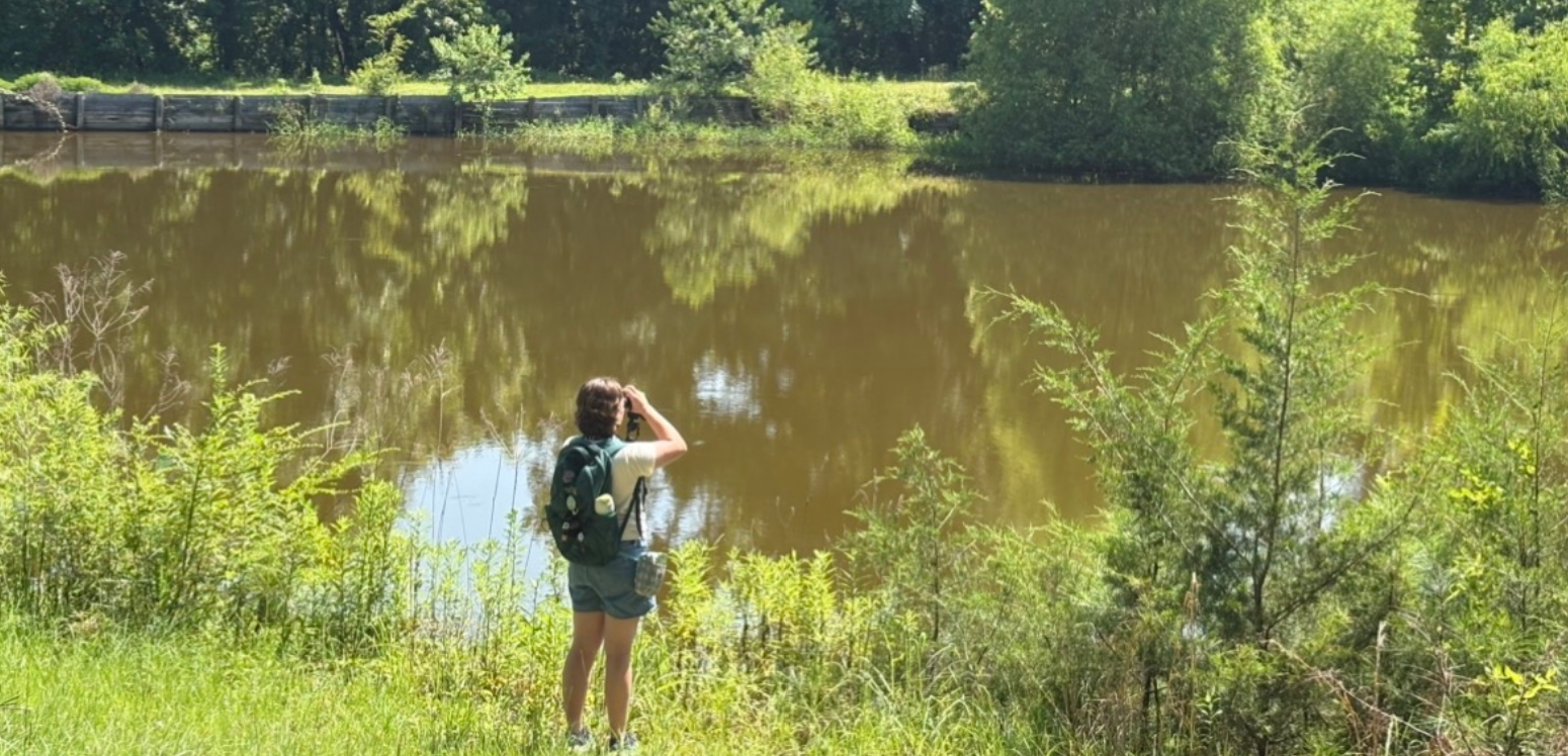 AG stands facing away from the camera and towards a lake, looking through binoculars.
