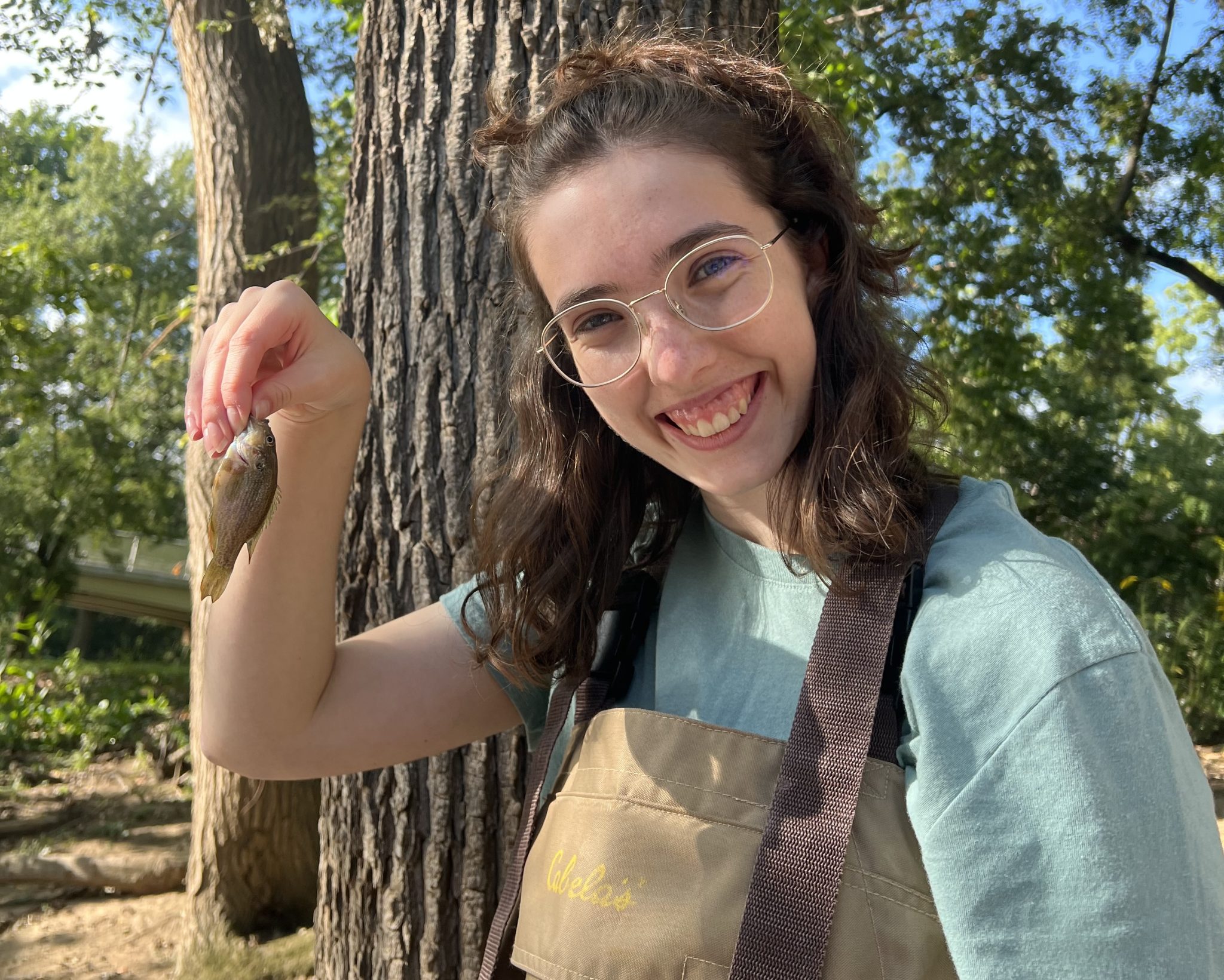 A woman with brown hair and glasses wearing a blue shirt and brown waders smiles while holding a small fish.